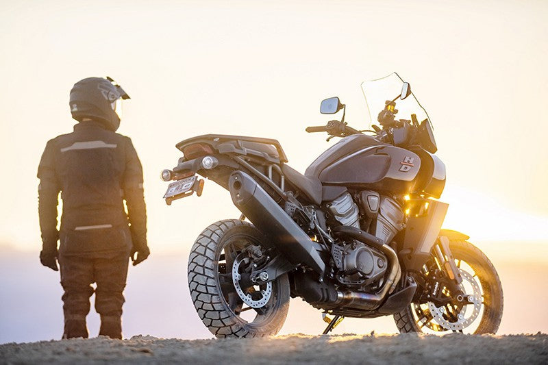 Mężczyzna motocyklista w kombinezonie motocyklowym i kasku stojący obok swojego motocykla turystycznego marki Harley-Davidson na tle zachodzącego słońca