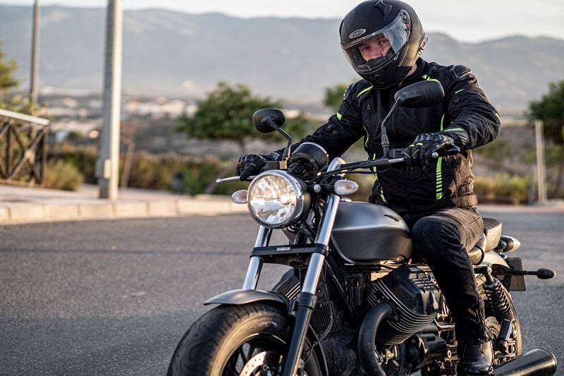Motocyklista w kurtce motocyklowej ozone i kasku, siedzący na motocyklu typu naked, znajduje się na asfaltowej drodze, w tle krzewy i góry