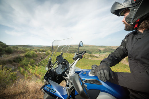 Motocyklista siedzący na motocyklu w kasku motocyklowym z podniesioną szczęką, patrzący w dal, w tle zielone pagórki