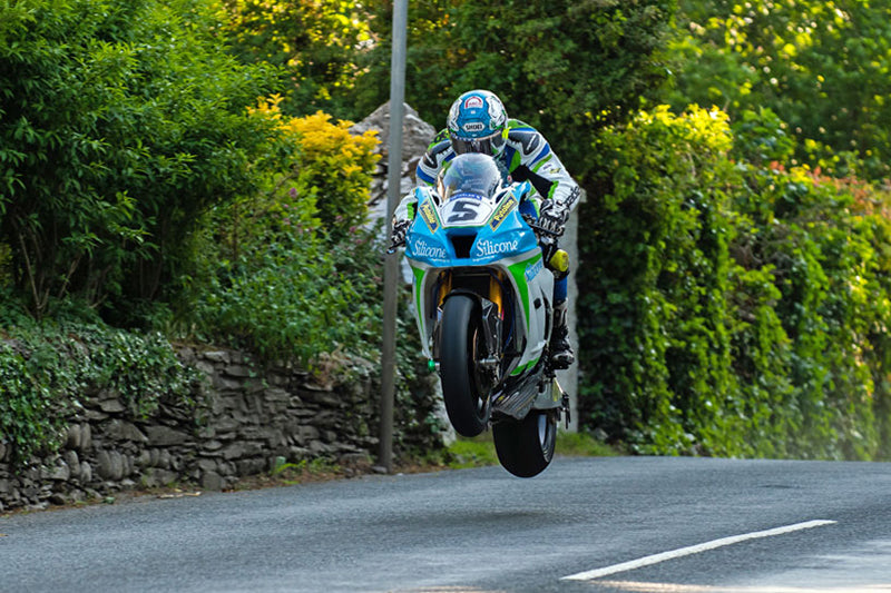 Motocyklista Dean Harrison podczas wyścigu, zdjęcie uchwycone w momencie wyskoku motocykla, w tle drzewa