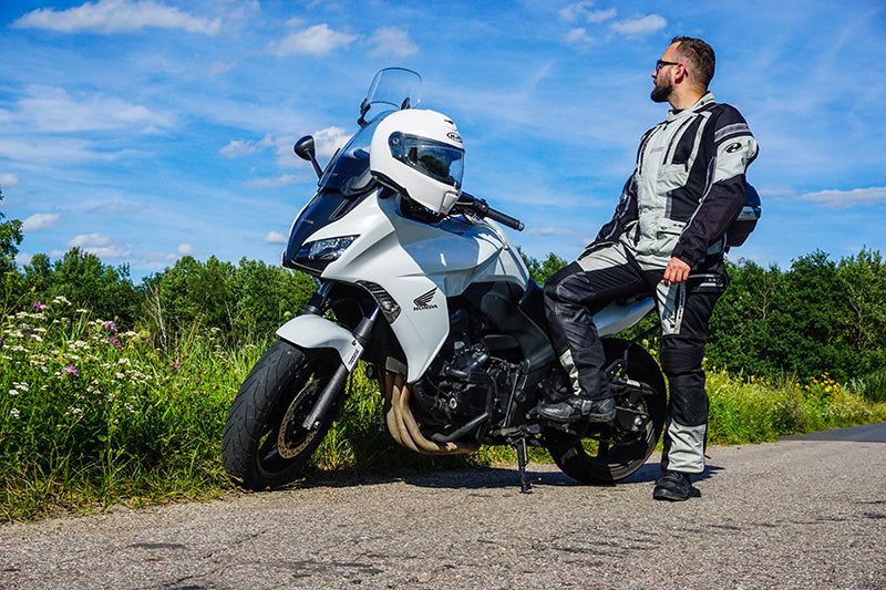 Motocyklista w kombinezonie motocyklowym i ciemnych okularach, pozujący przy swoim białym motocyklu, na kierownicy którego zawiesiony jest kask, w tle łąka i niebo