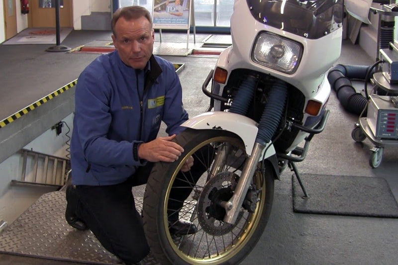 Pracownik w warsztacie samochodowym w niebieskiej kurtce kucający przy białym motocyklu, w tle kanał samochodowy