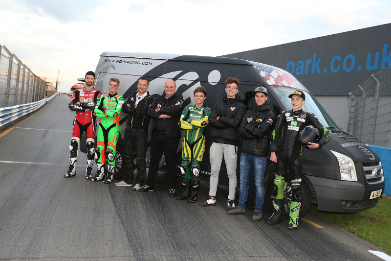 Grupa osób przed busem pozująca do zdjęcia, wśród nich motocykliści w kombinezonach motocyklowych