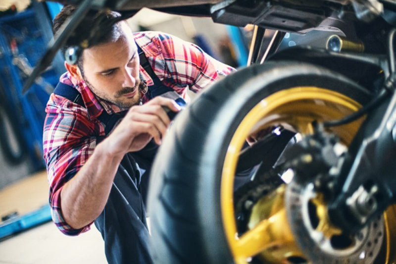 Mechanik z koszuli w kratę w warsztacie naprawia motocykl, widoczne koło motocykla z żółtą felgą