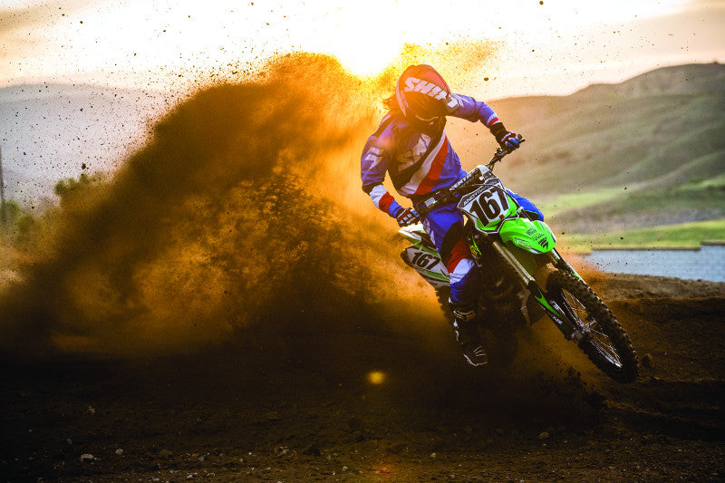 Motocyklista w offroadzie jadący po torze w kasku i kombinezonie motocyklowym, za nim kurz piachu i zachód słońca