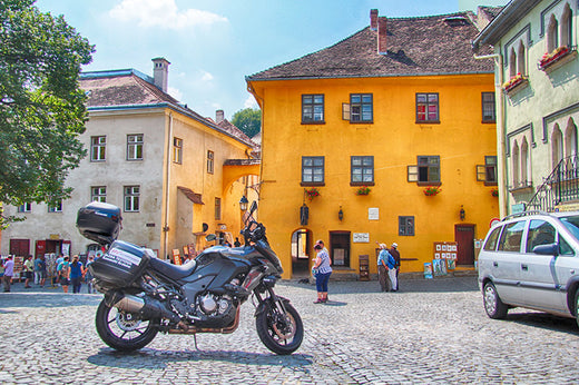 Motocykl zaparkowany na środku betonowego placu w słoneczny dzień, w tle żółty budynek i inne budynki oraz spacerujący ludzie i samochód
