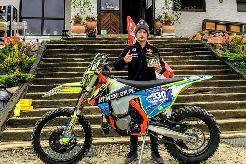 Franek Dubaniowski motocyklista enduro, wraz ze swoim motocyklem, z wyciągniętym kciukiem, w tle schody i budynek
