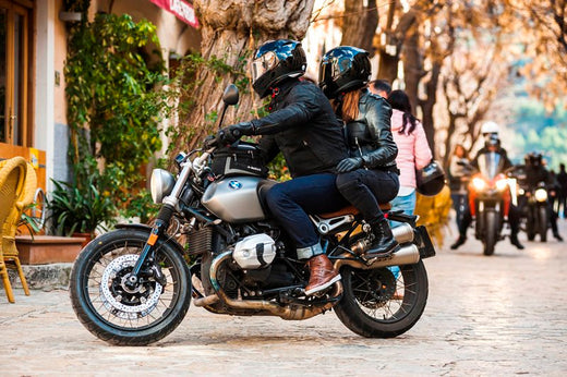 Para na motocyklu BMW w kaskach, ustawiona bokiem przy kawiarni, w tle inni motocykliści