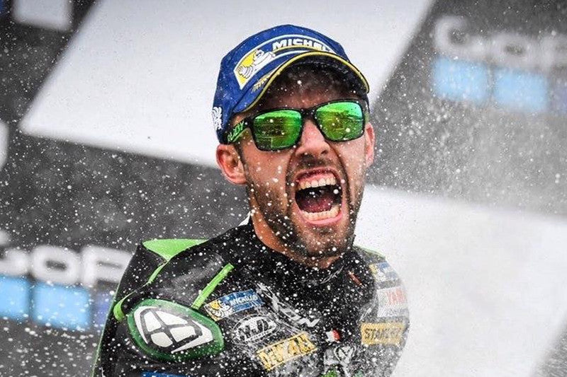 Jonas Folger podczas MotoGP Niemiec, cieszący się ze zwycięstwa, oblewany szampanem, w czapce i okularach przeciwsłonecznych