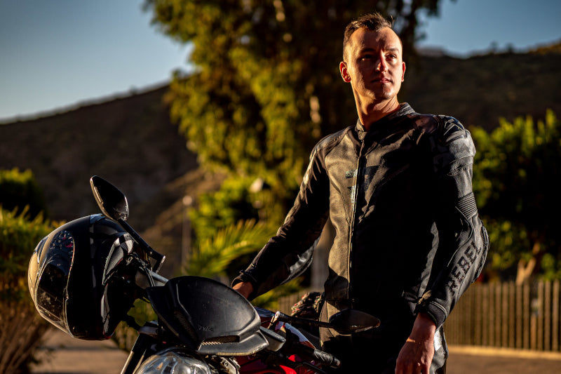 Motocyklista w czarnej skórzanej kurtce motocyklowej patrzy w dal, obok motocyklisty motocykl z kaskiem zawieszonym na kierownicy, wieczorowa pora, w tle drzewa