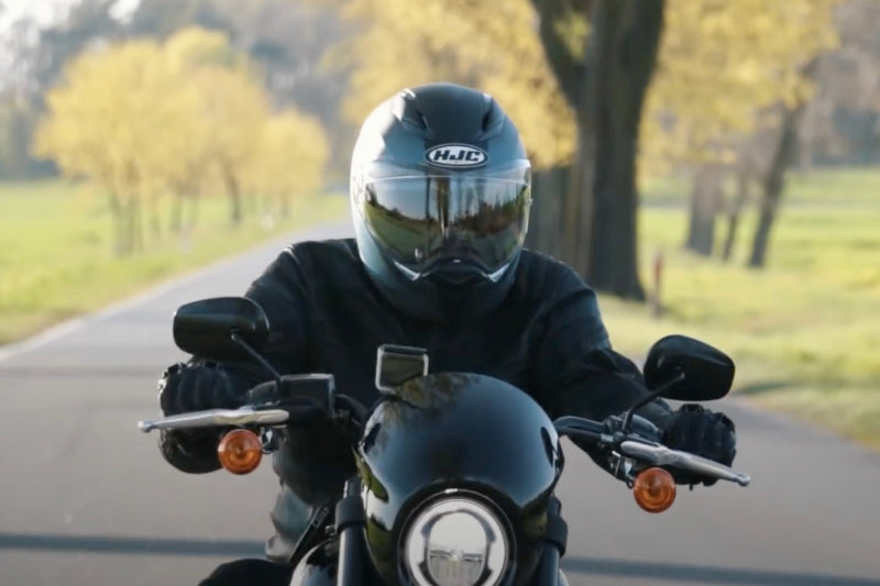 Motocyklista jadący na motocyklu w kasku HJC z rękoma na manetkach, w tle pozamiejska droga i drzewa