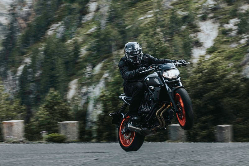 Motocyklista w czarnym kombinezonie motocyklowym i kasku HJC, robiący manewr motocyklowy na betonowym placu, w tle drzewa