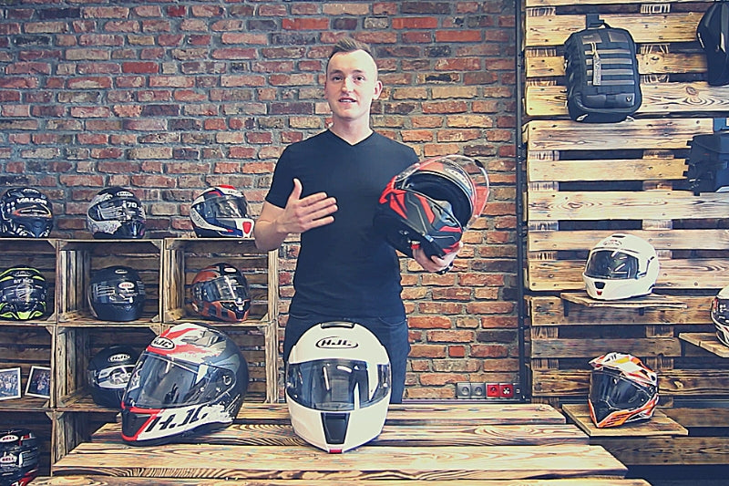 młody mężczyzna stoi w pomieszczeniu pełnym kasków motocyklowych, za nim ceglana ściana, a przed nim 3 modele kasków hjc