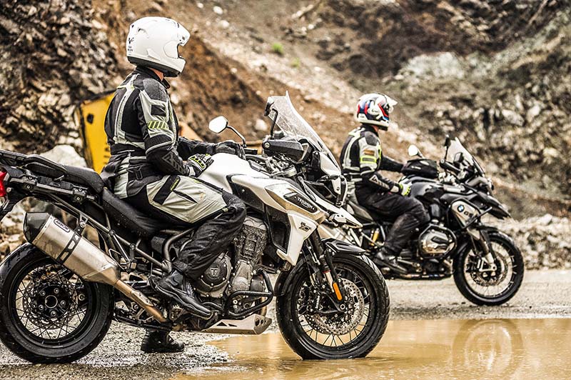 Dwóch motocyklistów w kaskach, siedzących na swoich motocyklach. Motocykle ustawione bokiem, na ziemi duża kałuża, po boku skarpy
