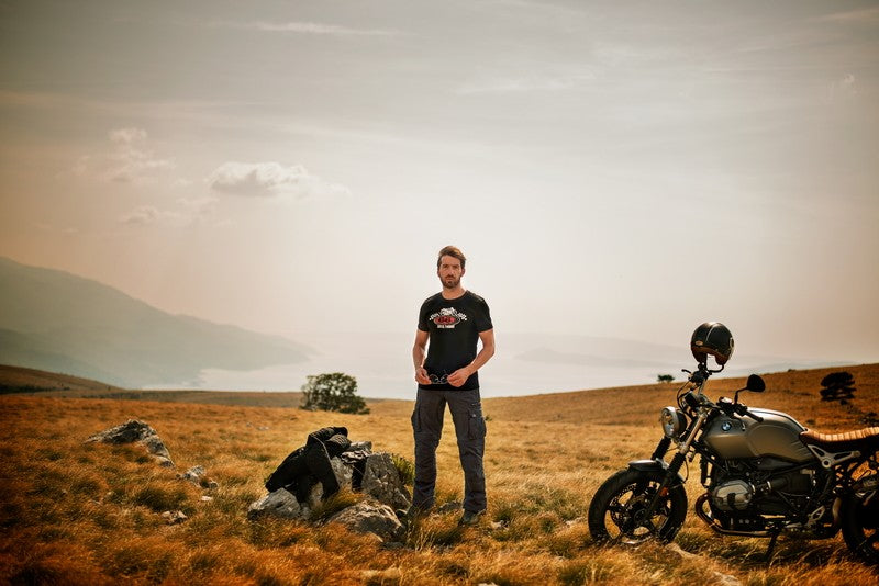 Mężczyzna w czarnej koszulce stojący na środku polany obok motocykla i kamienia, na którym leży jego skórzana kurtka, w tle jasne zamglone niebo i góry