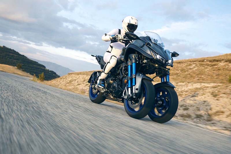 Dynamiczne zdjęcie motocyklisty na trójkołowym motocyklu yamaha, ubranym w biały kombinezon, zdjęcie robione od dołu, w tle droga jasne pagórki i niebo