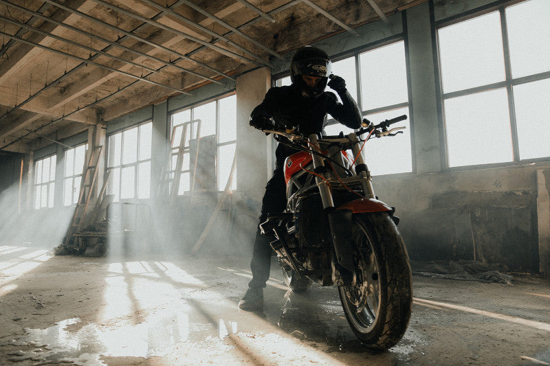Motocyklista w skórzanej kurtce i spodniach motocyklowych oraz kasku, siedzący na motocyklu typu naked w starej hali garażowej