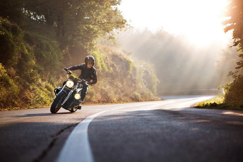 Motocyklista w drodze w kasku i skórzanym kombinezonie, w tle padające schodnie i dalsza część drogi oraz jesienny las