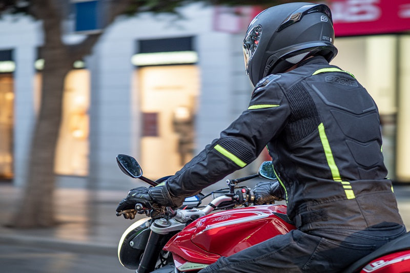 Motocyklista widoczny bokiem podczas jazdy, ubrany w kurtkę motocyklową ozone i kask motocyklowy, w tle witryny sklepowe 