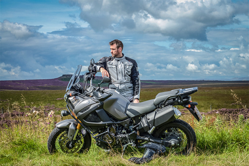 Motocyklista w kombinezonie motocyklowym RST Adventure stojący przy swoim motocykli turystycznym na polanie, w tle niebo i pagórki