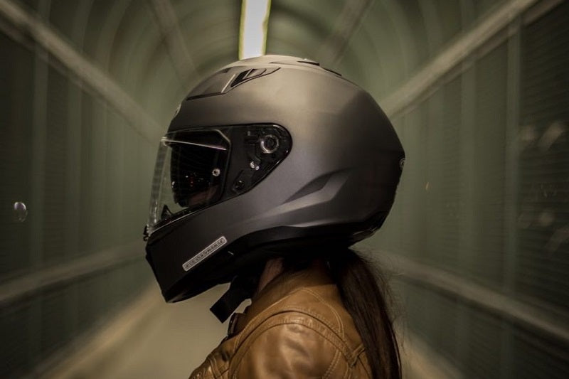 Głowa kobiety stojącej bokiem w ciemnym grafitowym kasku motocyklowym, w tle długi ciemny tunel