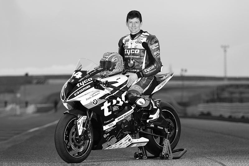 Czarno białe zdjęcie zmarłego motocyklisty. Dan Kneen siedzący na swoim motocyklu z uśmiechem, w tle droga.