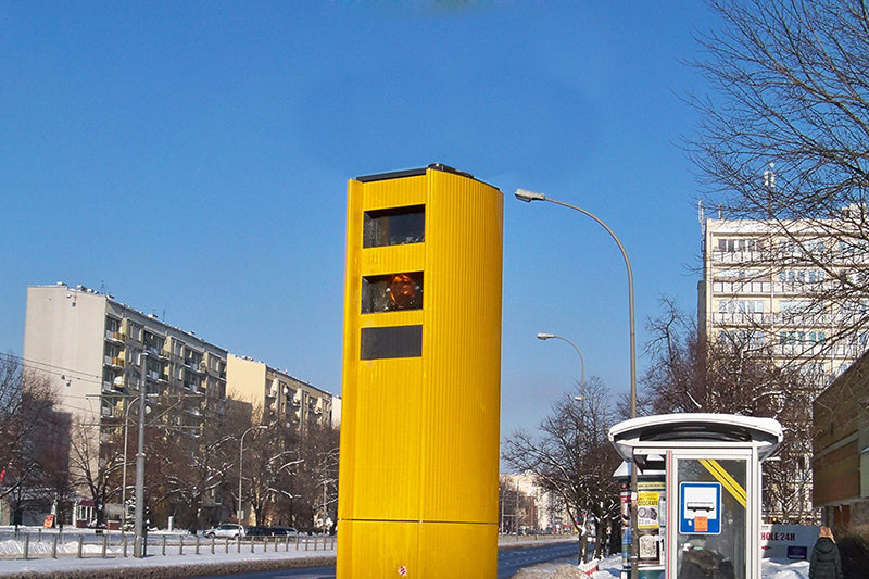 Żółty fotoradar w mieście stojący w pobliżu przystanku autobusowego, w tle bloki i jasne niebieskie niebo
