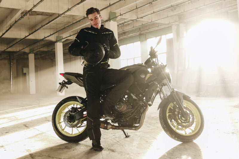 Motocyklista w kombinezonie motocyklowym, pozujący przy swoim motocyklu w hali garażowej, trzyma w dłoniach kask motocyklowy