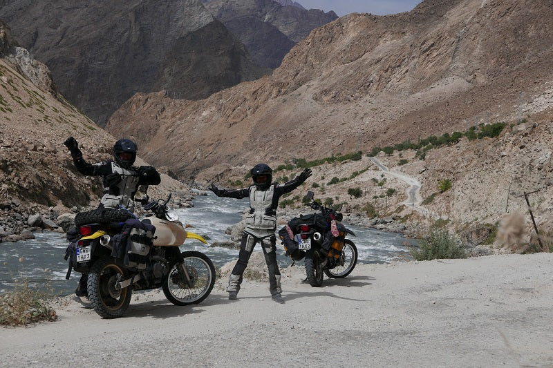 Para motocyklistów stojąca w kaskach motocyklowych i strojach obok swoich motocykli, pozdrawiając do zdjęcia, w tle góry i woda