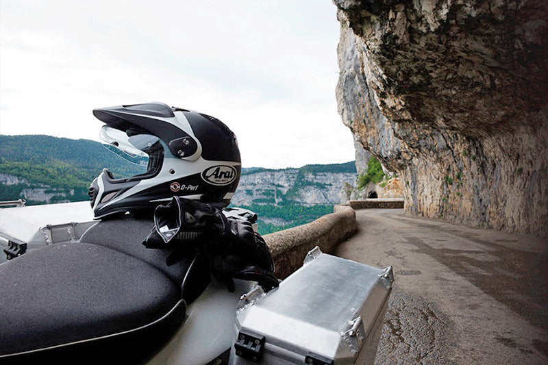 Kask motocyklowy typu adventure położony na motocyklu turystycznym wyposażonym w kufry motocyklowe, w tle skalna droga