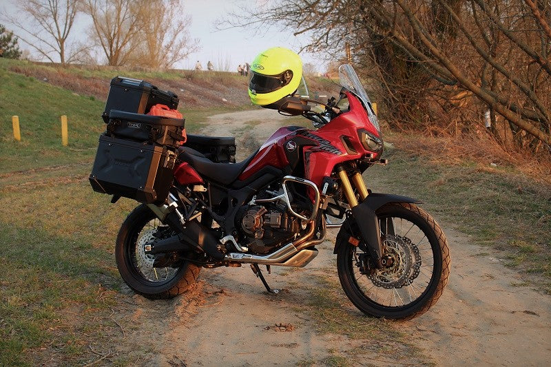 Czerwony motocykl z kuframi na bagażniku i żółtym kaskiem zawieszonym na kierownicy, stojący na leśnej drodze, obok drzewa i pagórki