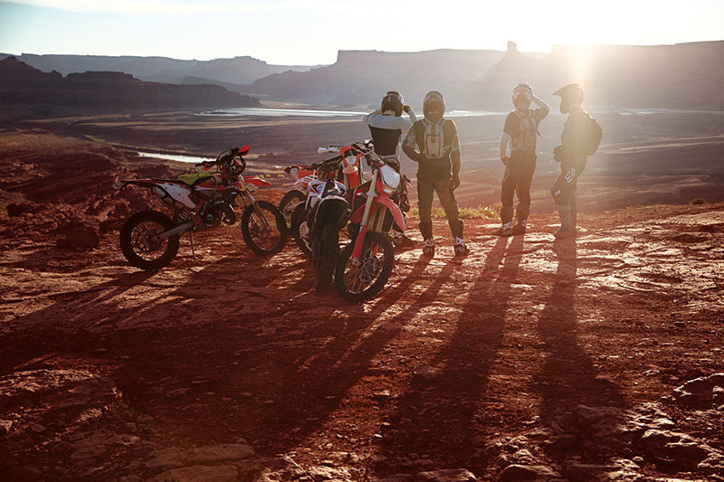 Grupa motocyklistów enduro stoi nad skalistym pustynnym urwiskiem ubrana w stroje Fox Legion, w tle zachodzące za skalnym horyzontem słońce