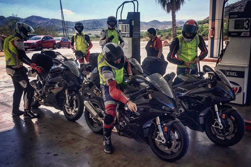 Grupa motocyklistów na swoich motocyklach marki BMW w kaskach i kamizelkach odblaskowych na stacji benzynowej, w tle góry i jasne niebo