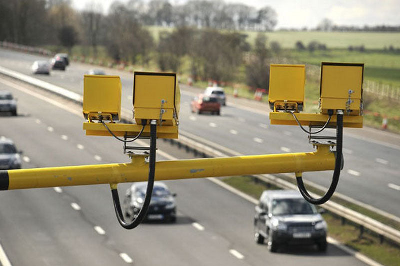 Dwie pary żółtych fotoradarów na poziomym słupie zawieszone and ulicą, w tle autostrada z jadącymi autami