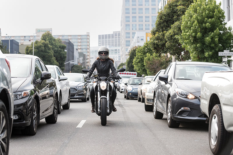 Motocyklista jadąca ulicą w kasku, okularach przeciwsłonecznych i skórzanej kurtce, po bokach jadące samochody, w tle wieżowce i drzewa