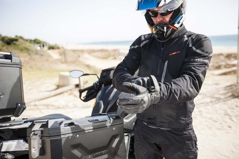 Motocyklista w kasku i okularach przeciwsłonecznych zakładający rękawice motocyklowe, obok niego kufry przyczepione do motocykla, w tle piaszczysta plaża