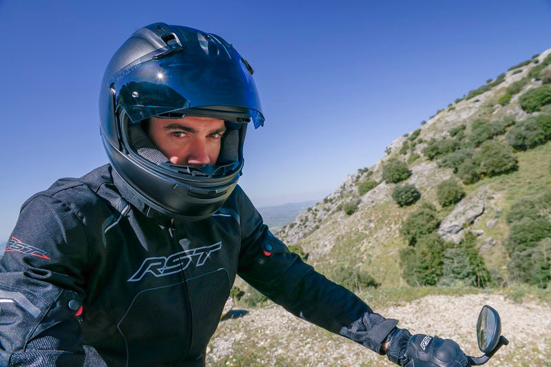 Motocyklista w kurtce motocyklowej RST i kasku motocyklowym z otwartą szybką, patrzący w obiektyw, siedząc na motocyklu, w tle niebo i pagórki