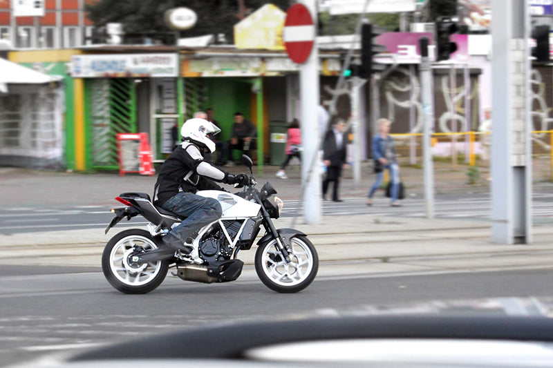 Motocyklista na białym motocyklu na skrzyżowaniu w mieście, skręcający w ulicę. Ubrany w biały kask. W tle witryny sklepowe i ludzie przechodzący przez pasy.