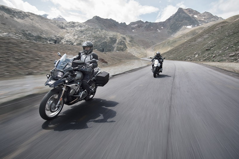 Dwaj motocykliści w kaskach na motocyklach turystycznych, jadący jeden za drugim po asfalcie, w tle górskie tereny i niebo