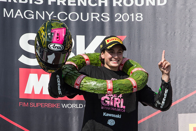 Kobieta, Ana Carrasco, z tytułem mistrza świata w wyścigach motocyklowych, cieszy się z wygranej, stoi w czapce z daszkiem, trzyma kask i wyciąga palec wskazujący do góry