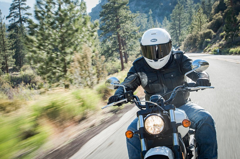 Motocyklista w białym kasku Bell i w skórzanej kurtce motocyklowej, jedzie drogą w górach na motocyklu.