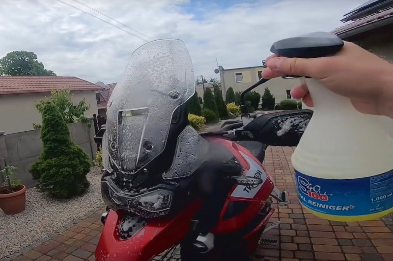 Motocykl marki Triumph myty na podjeździe przed domem, ręka trzyma przed nim środek do czyszczenia i pielęgnacji motocykla