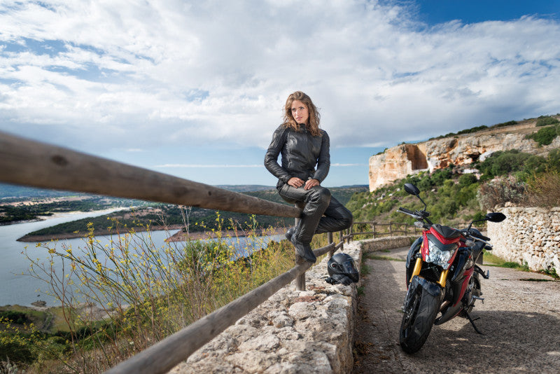 motocyklistka siedzi na barierce przy górskiej drodze, obok stoi jej czerwony motockyl