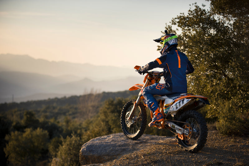 Motocyklista offroadowy na zboczu skarpu, siedzi na motocyklu w kasku motocyklowym i stroju marki fox, patrzący w niebo