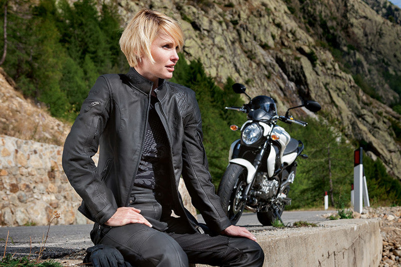 Motocyklistka blondynka w czarnej skórzanej kurtce motocyklowej, siedząca na murku na poboczu drogi, obok niej zaparkowany motocykl