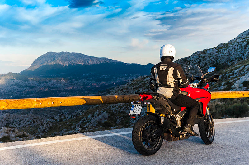 Motocyklista siedzący na czerwonym motocyklu, w kurtce motocyklowej i kasku, znajduje się on w punkcie widokowym w górach, w tle jasne niebo i góry