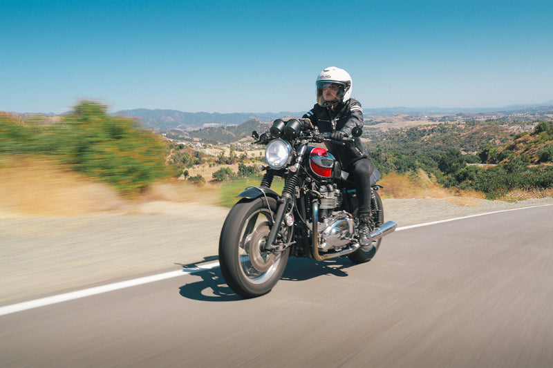 motocyklistka jadąca drogą poza miastem, ubrana w skórzaną kurkę, czarne jeansy a na głowie ma biały kask motocyklowy typu orzeszek - HJC i30