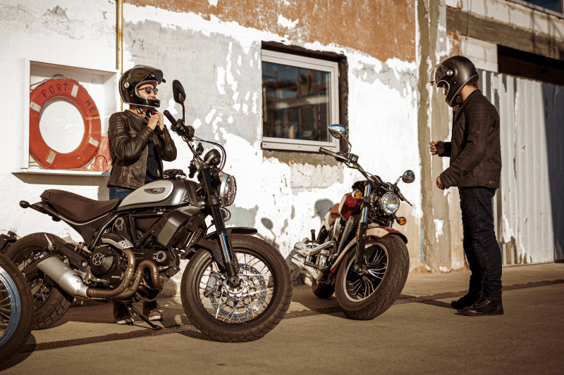 Para motocyklistów na postoju, motocyklistka poprawia kask motocyklowy, obok nich dwa motocyklowe i stara ściana budynku
