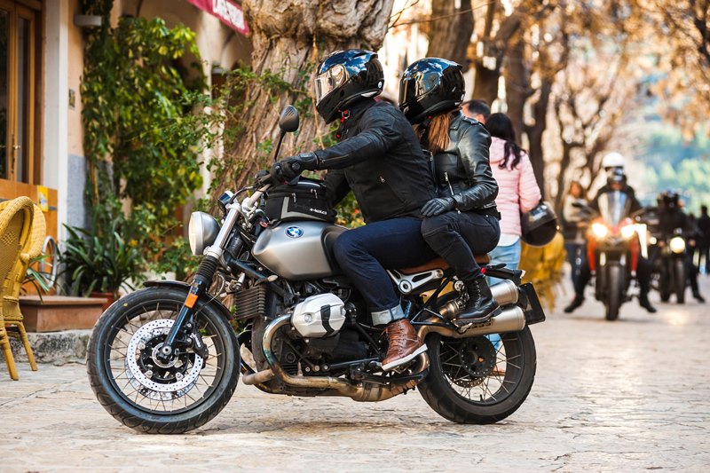 Para na motocyklu BMW w kaskach, ustawiona bokiem przy kawiarni, w tle inni motocykliści 