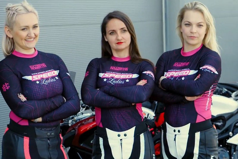 3 dziewczyny w kombinezonach i koszulkach Speed Ladies w tle motocykle